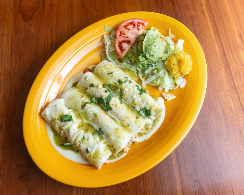 Three green enchiladas with sour cream, lettuce, guacamole, tomato, and corn on a yellow plate.
