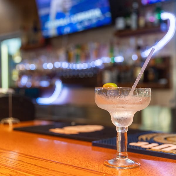 A frosty margarita with a lime wedge and straw sits on a wooden bar counter.