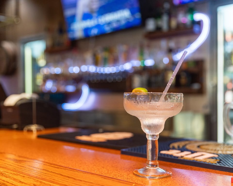 A frosty margarita with a lime wedge and straw sits on a wooden bar counter.