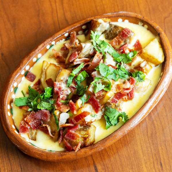 Melted cheese dish with bacon, potatoes, and cilantro in an oval clay dish on a wooden table.