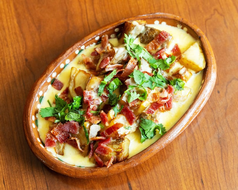 Melted cheese dish with bacon, potatoes, and cilantro in an oval clay dish on a wooden table.