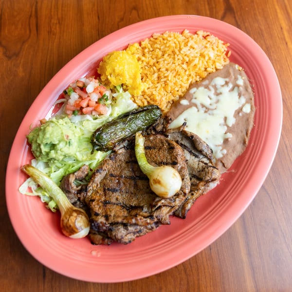 Grilled steak with rice, refried beans, guacamole, pico de gallo, and grilled onions on a pink plate.