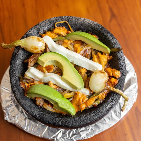 Molcajete dish with meat, cheese, avocado, and onions in a black stone bowl on a wooden table.