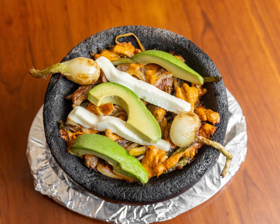 Molcajete dish with meat, cheese, avocado, and onions in a black stone bowl on a wooden table.