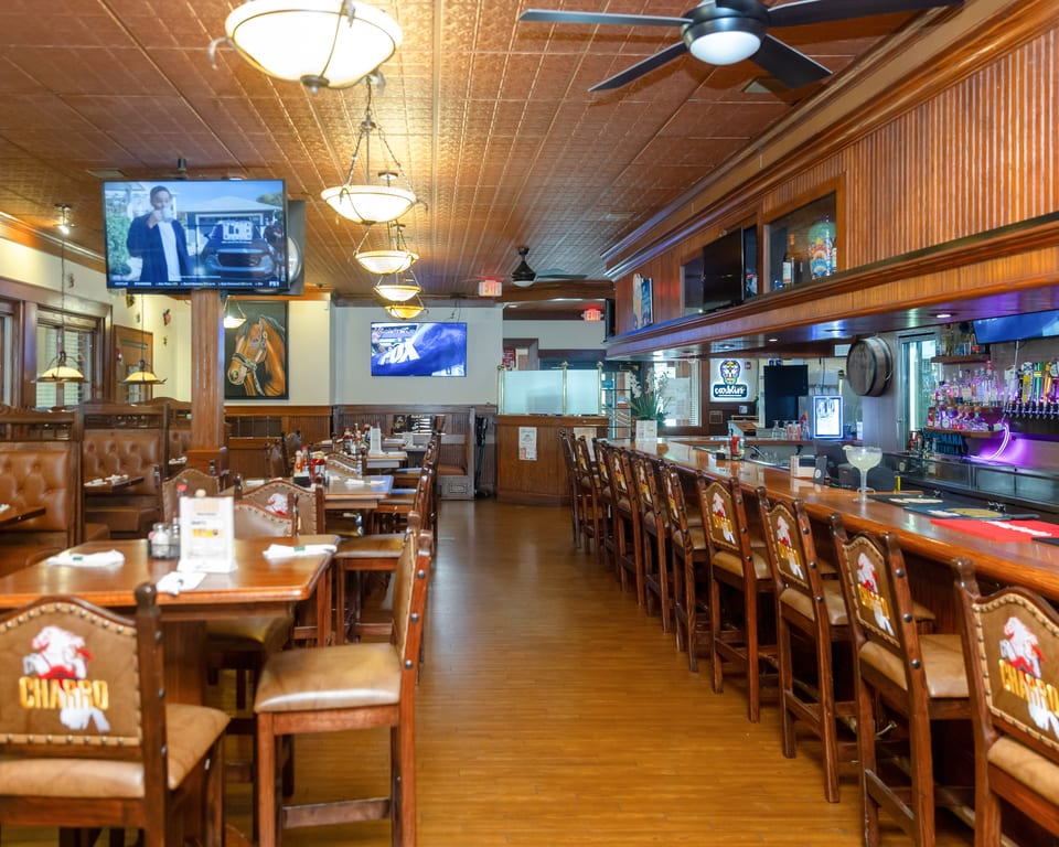 Spacious restaurant interior with wooden tables, brown booths, a long bar with stools, and TVs on the walls.