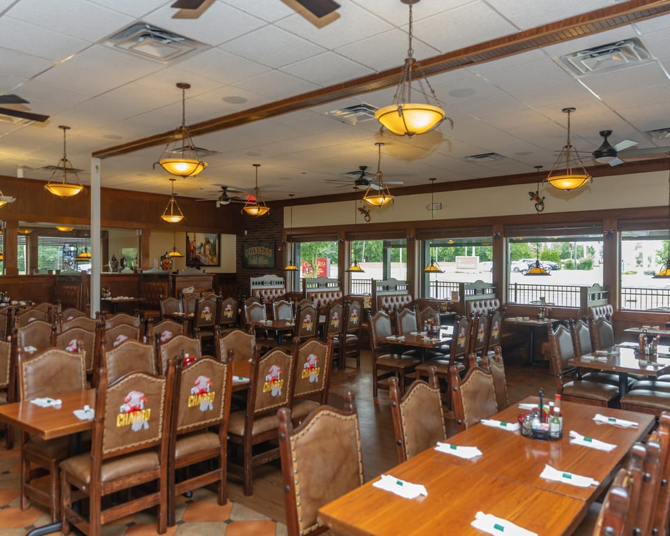 Spacious restaurant dining room with many empty tables and chairs, featuring large windows and warm lighting.