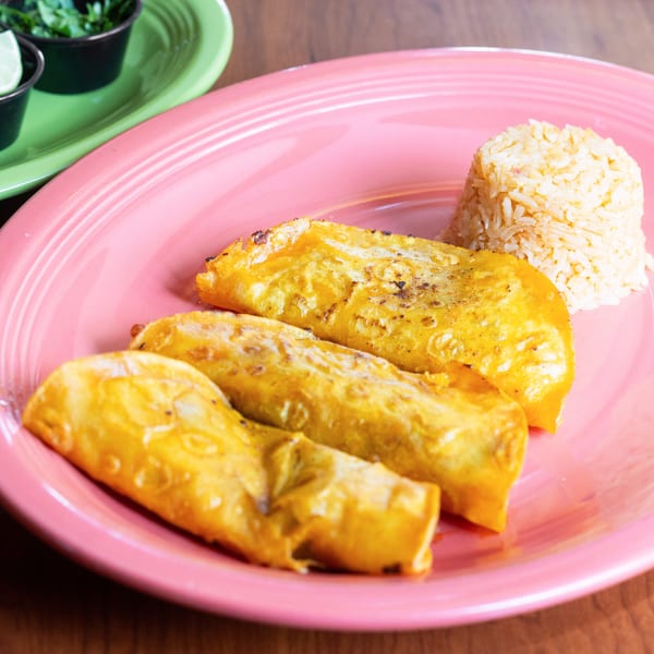 Three golden-brown enchiladas and a mound of rice on a pink plate, with lime and cilantro in the background.