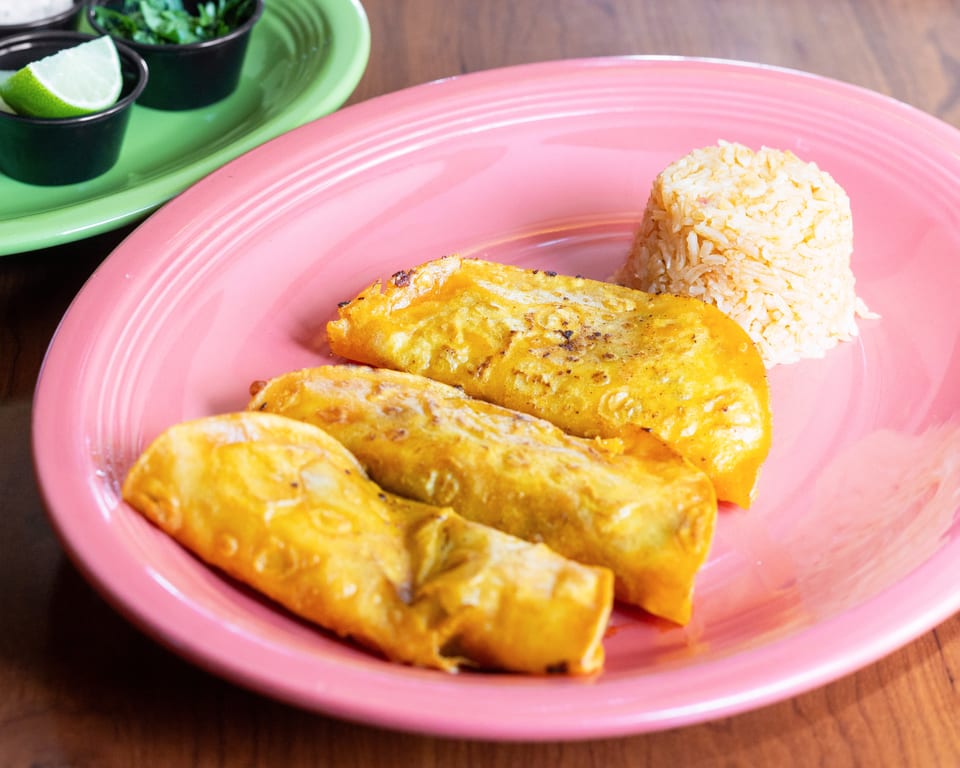 Three golden-brown enchiladas and a mound of rice on a pink plate, with lime and cilantro in the background.