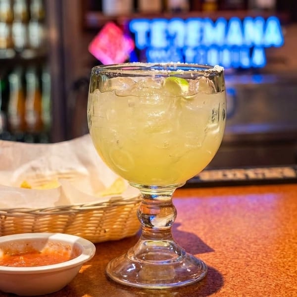 Margarita with lime, chips, and salsa on a bar counter with a neon sign in the background.