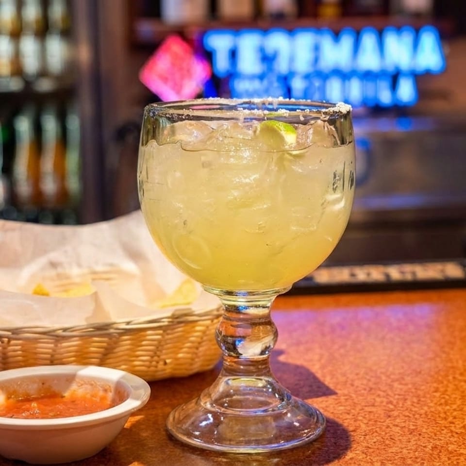 Margarita with lime, chips, and salsa on a bar counter with a neon sign in the background.